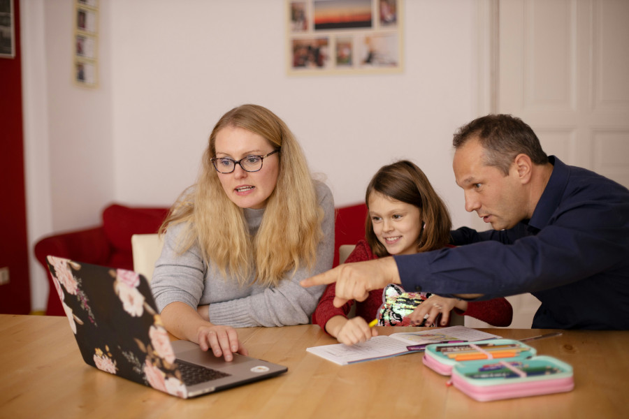 Kobieta w okularach korzysta z laptopa, siedząc przy stole z mężczyzną i dziewczynką; wspólnie omawiają zadania szkolne.